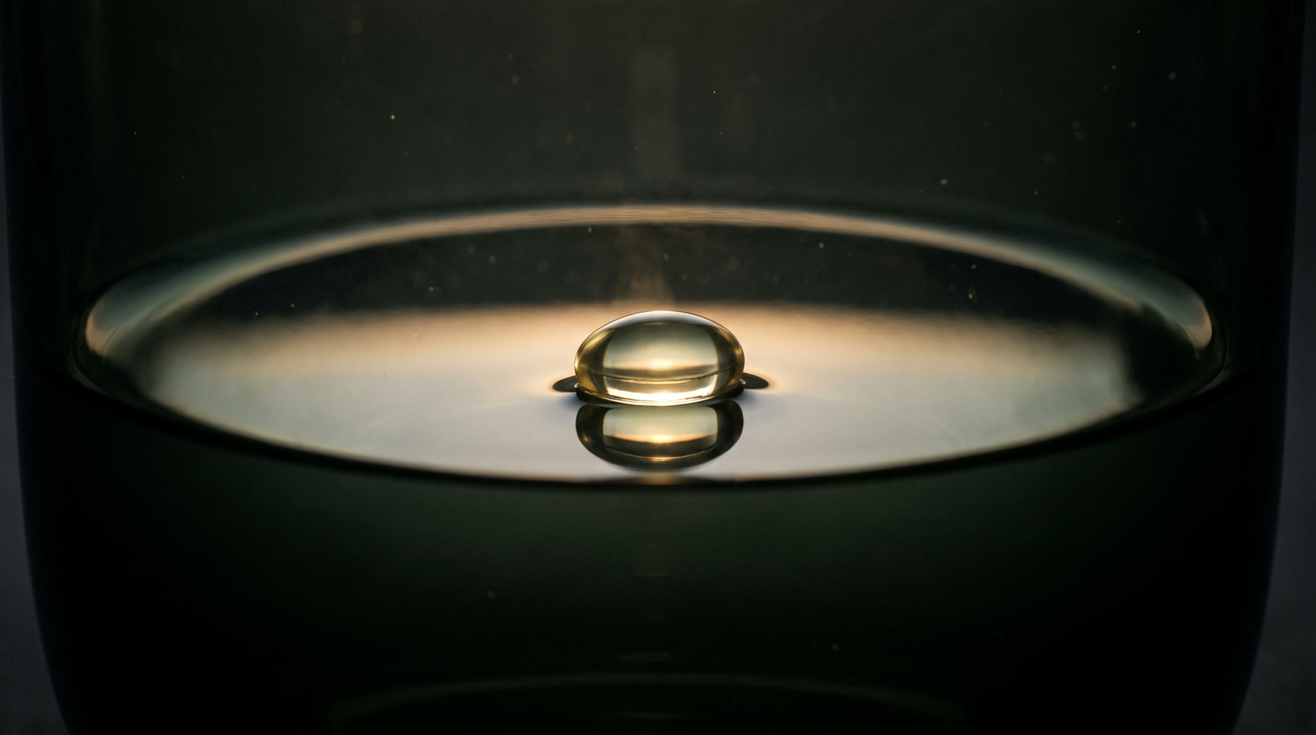 A single clear softgel of pale gold oil resting on still dark water, lit by one thin line of light.